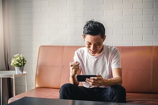 Junger Mann sitzt auf der Couch mit dem Handy in der Hand und gewinnt ein Handyspiel. 